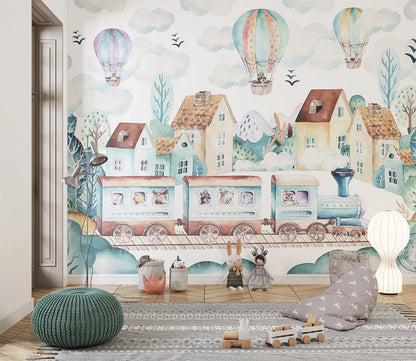 Watercolor illustration features a picturesque village with charming houses and lush trees, set against a backdrop of snowy mountains. A whimsical train with animal passengers rolls along the tracks, while colorful hot air balloons float serenely in the sky. The scene radiates a dreamy, adventurous atmosphere filled with soft pastel tones.