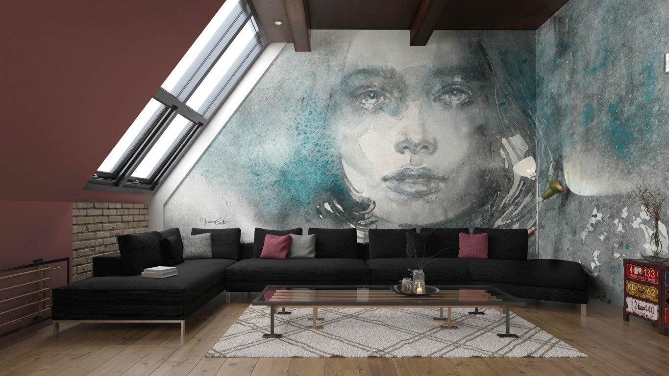 Adriana - Custom Wallpaper Mural l Wallab Egypt. A stylish loft living room featuring a striking wall mural of a woman's ethereal portrait in watercolor, blending muted tones with splashes of teal. The space includes a large black sectional sofa adorned with grey and pink throw pillows, a modern glass coffee table, and a patterned rug, all under a slanted ceiling with skylights that bring in natural light.