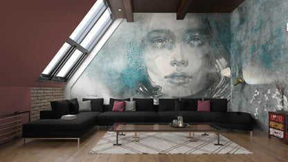 Adriana - Custom Wallpaper Mural l Wallab Egypt. A stylish loft living room featuring a striking wall mural of a woman's ethereal portrait in watercolor, blending muted tones with splashes of teal. The space includes a large black sectional sofa adorned with grey and pink throw pillows, a modern glass coffee table, and a patterned rug, all under a slanted ceiling with skylights that bring in natural light.